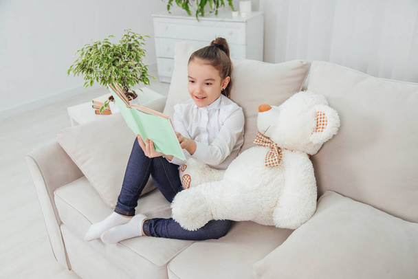 Cute little girl sitting on the sofa with teddy-bear near her, reading fairy-tale, pointing her finger at something in the book, smiling. - Fotoğraf, Görsel