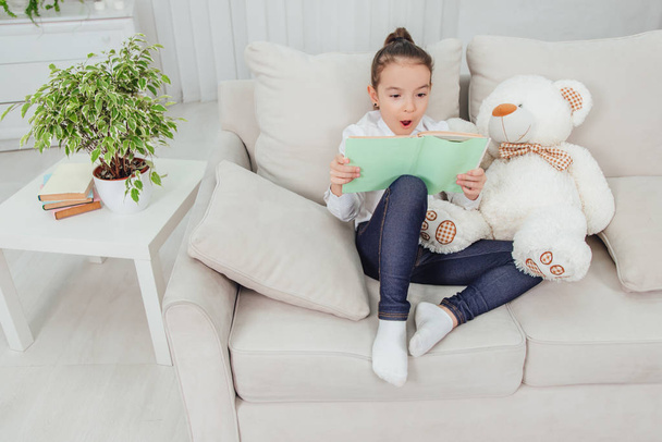 Cute little girl sitting on the sofa with teddy-bear near her, reading fairy-tale, opening her mouth in excitement. - Photo, Image