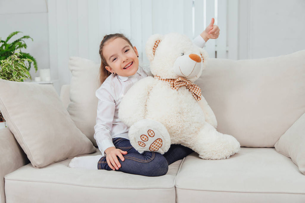 Smiling little kid is sitting on the sofa with white plush bear, looking at the camera, giving her thumb up. - Foto, afbeelding