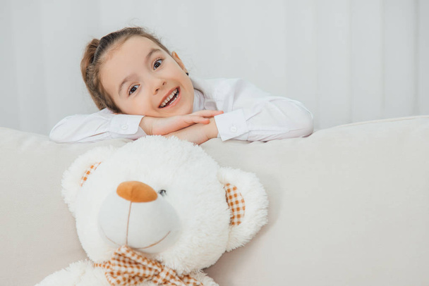 Big white soft bear is on the sofa. Girl is standing behind the sofa, leaning over its back, folding her hands, looking at the camera, smiling pleasantly. - Фото, изображение