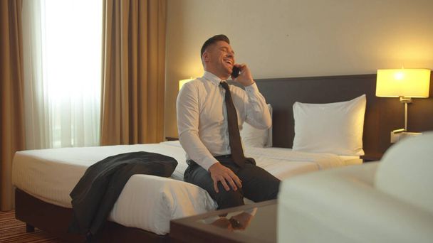 Man in shirt and tie sit on the bed in hotel and talk on phone with laugh  - Foto, Bild