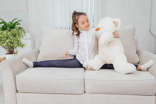 Sporty flexible girl is sitting on the sofa, making split, hugging white teddy-bear. - Photo, Image