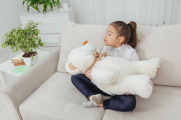 Cute small girl sitting on the sofa in lotos position, lulling her white teddy-bear. - 写真・画像