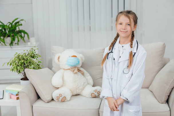 Vrij zelfverzekerd klein meisje in doktersjas, stethoscoop staand, kijkend naar de camera, lachend. - Foto, afbeelding