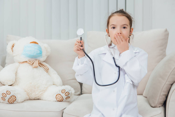 Little girl in medical coat holding a stethoscope, covering mouth with her hand. White teddy-bear in medical mask is sitting near her. - Foto, Imagem