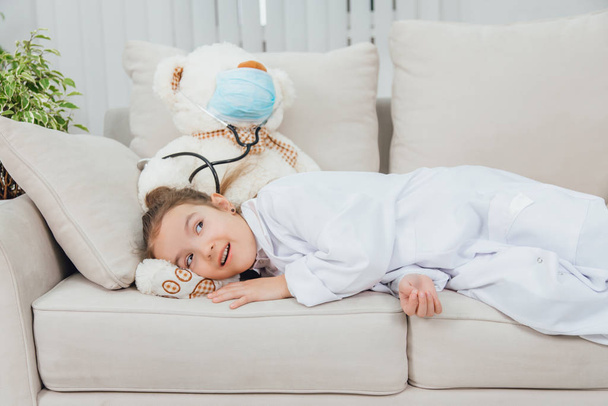 Little girl in medical coat is lying on the bears paw, looking lovely, smiling. White teddy-bear in medical mask and with stethoscope. - Fotó, kép