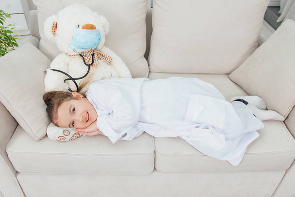 Little girl in medical coat is lying on the bears paw, looking lovely, smiling. White teddy-bear in medical mask and with stethoscope. - Фото, зображення