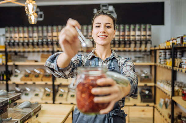 Young Mixed Race Woman Owner Selling Superfoods in Zero Waste Shop. Lots of Healthy Food in Glass Bottles on Stand in Grocery Store. No plastic Conscious Minimalism Vegan Lifestyle Concept. Copy space - Fotoğraf, Görsel