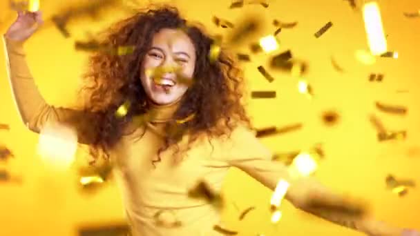 Excited girl dancing, applauding, having fun, rejoices over confetti rain in yellow studio. Concept of Christmas, New Year, happiness, party, winning - Video