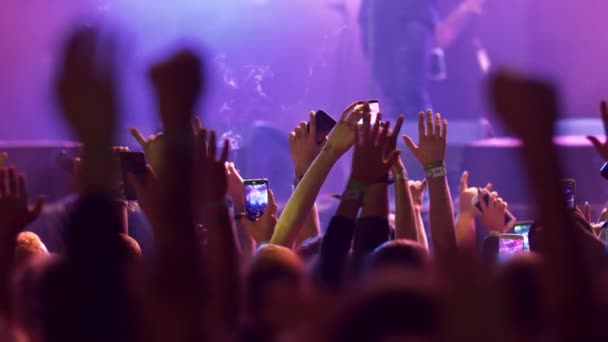 Az emberek feltartott karokkal állnak, szarvgesztusokat mutatnak, okostelefonjaikkal lövöldöznek, miközben egy dalt hallgatnak egy rock koncerten. A színpad lila világítással van megvilágítva. 4k - Felvétel, videó