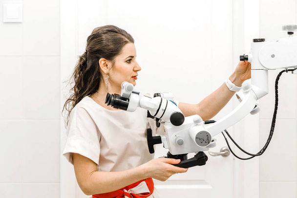Приваблива молода жінка-стоматолог у білому костюмі використовує мікроскоп на роботі
 - Фото, зображення