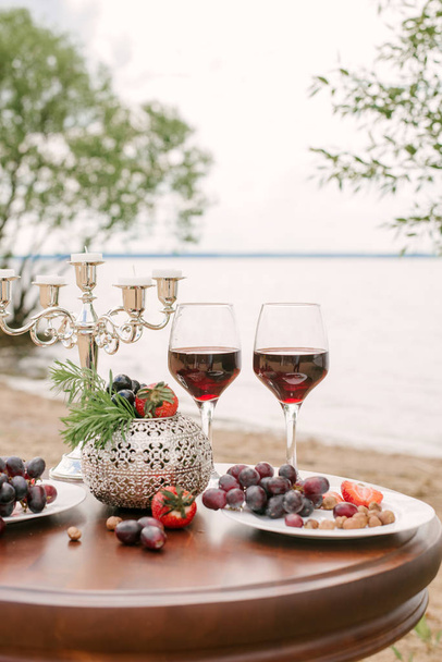 Ρομαντικό δείπνο του Αγίου Βαλεντίνου στην παραλία: δύο ποτήρια κόκκινο κρασί, φρέσκα φρούτα και ένα κηροπήγιο σε ένα ξύλινο τραπέζι - Φωτογραφία, εικόνα