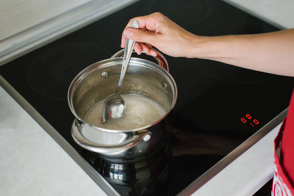 Женская рука перемешивает жидкость сахарного сиропа в кастрюле на электрической плите столовой ложкой
 - Фото, изображение