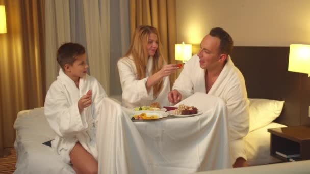 両親と息子はベッドに座ってホテルの部屋で食べ物を食べる - 映像、動画