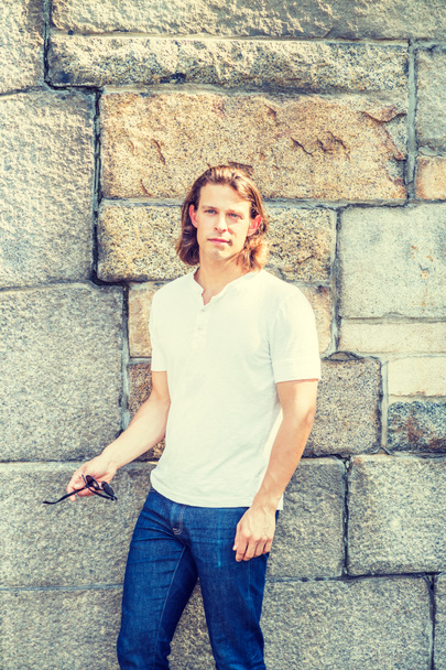 Portrait of Young Handsome American Man with long dark brown hair in New York City, wearing white short sleeve shirt, blue jeans, hand holding sunglasses, standing by rocks wall in summer under sun - Photo, Image