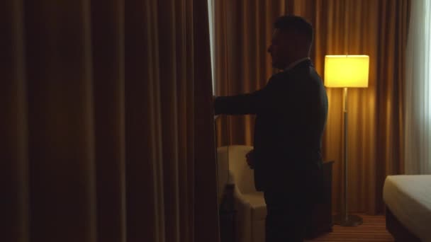 Man in jacket move curtains to look out the window in room of the hotel - Video