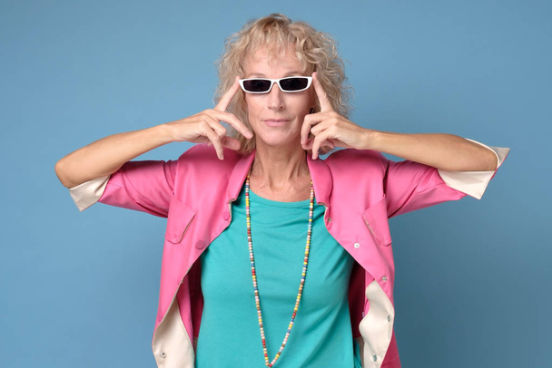Senior woman wearing stylish sunglasses and smiling at camera on blue wall. - Фото, изображение