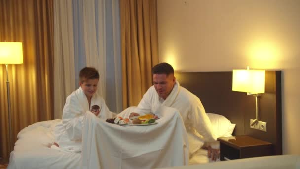 Πατέρας και γιος κάθονται στο κρεβάτι στο δωμάτιο του ξενοδοχείου και τρώνε το φαγητό - Πλάνα, βίντεο