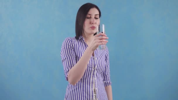 młoda kobieta z przyjemnością pije czystą, filtrowaną wodę ze szklanki stojącej na niebieskim tle - Materiał filmowy, wideo