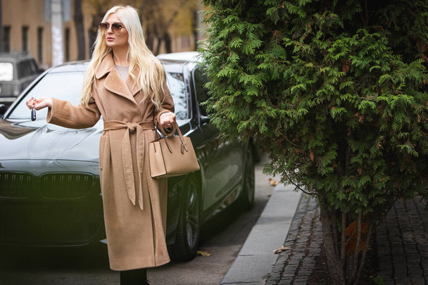 Young beautiful blond woman in an autumn coat stands on a city street. Soft focus. Fashionable concept. - 写真・画像