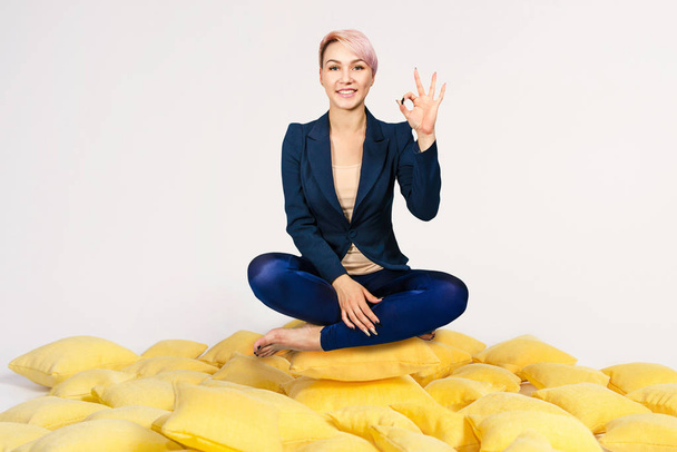Joven hermosa chica se sienta en una pose de loto en un montón de almohadas amarillas y muestra signo ok, sobre un fondo blanco
. - Foto, imagen