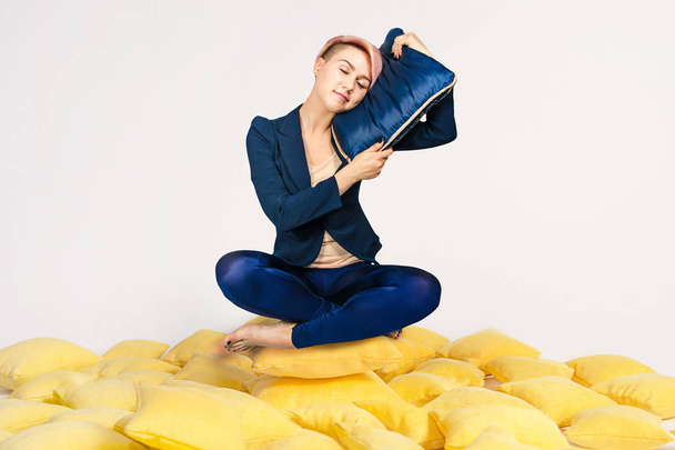 Young beautiful girl sits in a lotus pose on a pile of yellow pillows and sleeps on a blue pillow on a white background. - Photo, image