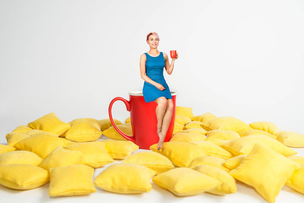 Young beautiful girl in blue dress sits on a huge cup in a yellow pillows and holds cup of coffee on a white background. - Fotoğraf, Görsel