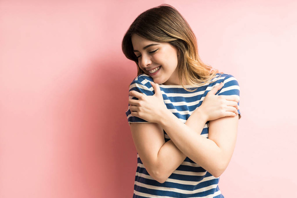Charming happy young woman hugging herself while isolated against pink background - Photo, Image