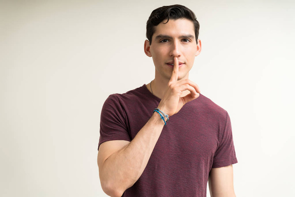 Πορτρέτο του ελκυστικού νεαρού άνδρα gesturing σιωπή με το δάχτυλο στα χείλη, ενώ στέκεται σε απλό φόντο - Φωτογραφία, εικόνα