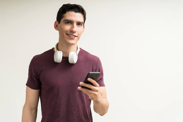 Usmívající se pohledný běloch se sluchátky držící smartphone, zatímco stojí na bílém pozadí - Fotografie, Obrázek