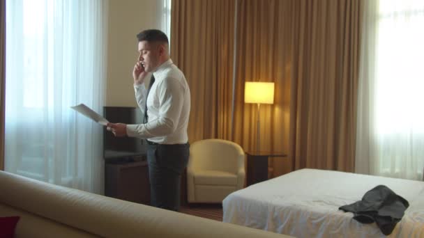Άντρας με έγγραφα στα χέρια μπαίνει στο δωμάτιο του ξενοδοχείου, μιλάει στο τηλέφωνο πολύ συναισθηματικά - Πλάνα, βίντεο