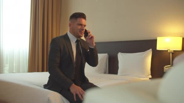 Мужчина в костюме сидит на кровати и разговаривает по телефону в отеле
 - Кадры, видео