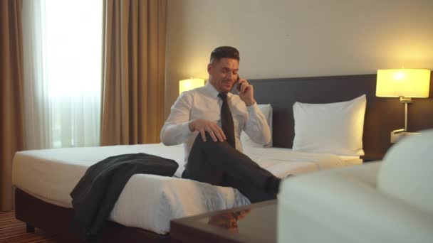 Мужчина в рубашке и галстуке сидит на кровати в отеле, разговаривает по телефону и снимает обувь
 - Кадры, видео