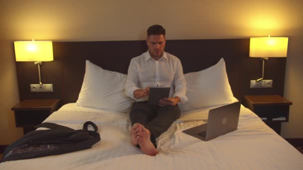 Obchodník v kalhotách sedí na posteli s tabletou v pokoji hotelu - Záběry, video