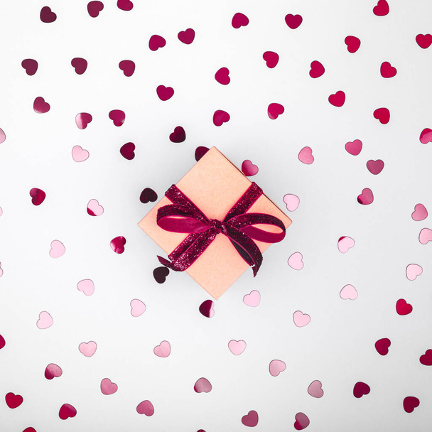 Caja artesanal con lazo de cinta de terciopelo rojo oscuro y confeti de corazón brillante. Día de San Valentín y concepto de envoltura ecológico. Fondo de diseño plano minimalista de moda
 - Foto, imagen