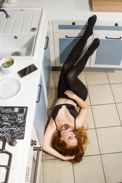 Rudowłosa dziewczyna leży na posadzce przy piecu w nowoczesnej kuchni - Zdjęcie, obraz