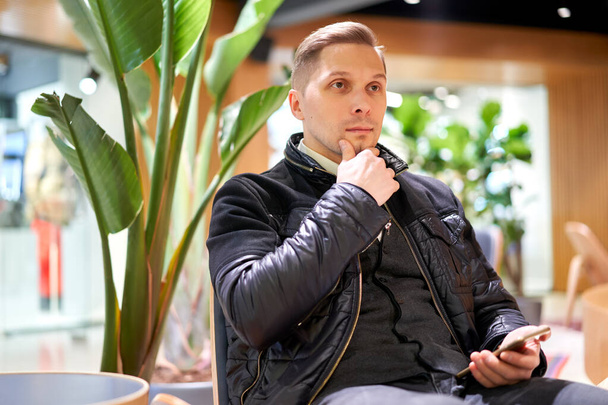 Male in leather jacket with smartphone looking at side while sitting on chair in store against green plant - Fotografie, Obrázek