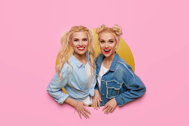 Image of two young happy shoked girlfriends women standing over pink background. Twins peep out of a hole in the background. Big Discount Season - Photo, Image