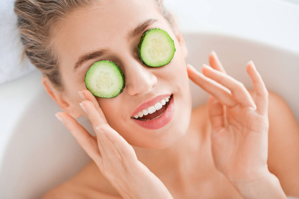 Beautiful young woman with cucumber slices lying in bathroom - Фото, изображение