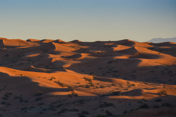 Güneş doğarken çöl, koyu turuncu renkli kumlu gölgeleri ortaya çıkarır ve Birleşik Arap Emirlikleri 'ndeki Ras al Khaimah' da dalgalanan veya dalgalanan tepeler üzerinde büyük bir çöl manzarası oluşturur.. - Fotoğraf, Görsel