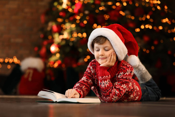 Little boy in Santa Claus cap reading book near Christmas tree a - Fotó, kép