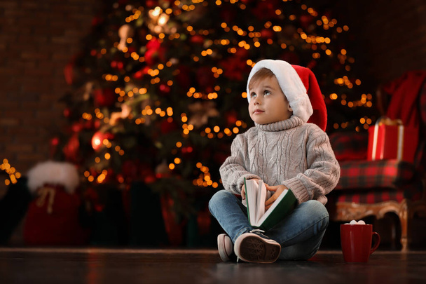 Little boy in Santa Claus cap with book near Christmas tree at h - Fotoğraf, Görsel