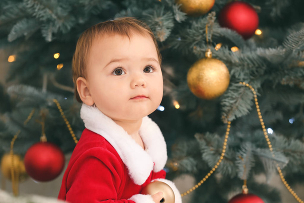Симпатичный малыш в костюме Санта-Клауса возле рождественской елки дома
 - Фото, изображение