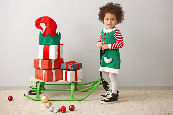Little girl in costume of elf, with Christmas gifts on sledge near light wall - Photo, image