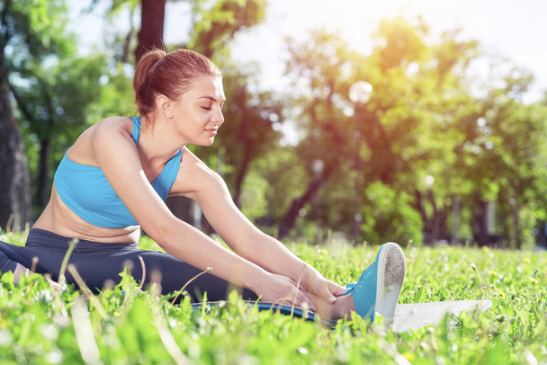 Attractive girl in sportswear doing yoga in park. Young woman sitting in yoga pose on green grass. Sport training outdoor at sunny summer day. Morning stretching exercises and healthy lifestyle. - Photo, Image