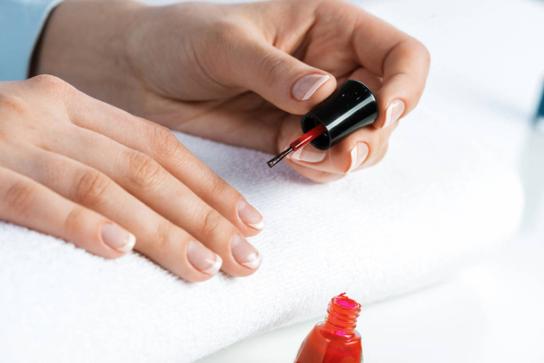 Woman giving herself elegant manicure at home. Closeup of beautiful female hand applying red nail polish at white table. Professional nail care and beautician procedure. Stylish nail art concept - Zdjęcie, obraz