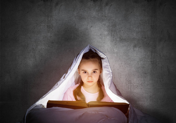 Little girl reading book in bed before going to sleep. Pretty child with flashlight hiding under blanket. Kid can not sleep at night. Portrait of beautiful girl in pajamas on background of grey wall. - Fotografie, Obrázek