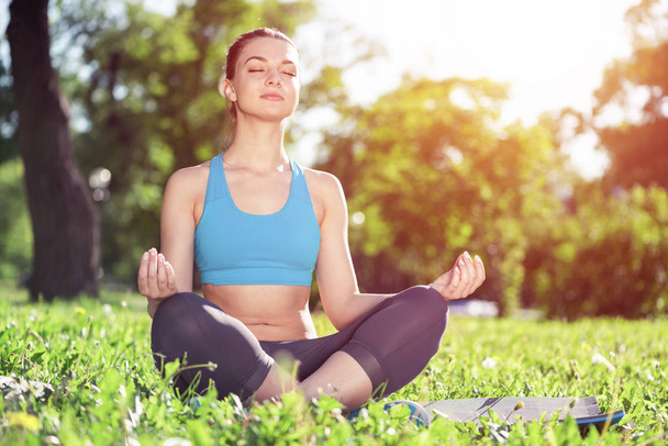 Girl meditates in lotus pose on green grass in park. Portrait of peaceful woman with closed eyes. Training and meditation outdoor at summer day. Harmony with nature and relaxation. Practicing of yoga. - Foto, immagini