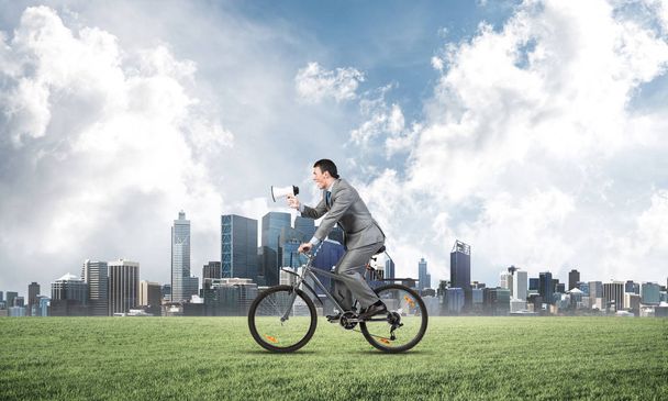 Businessman with megaphone on bike at sunny day. Marketing and advertising campaign. Manager in business suit riding bicycle on green meadow. Male cyclist on background of blue sky and city on horizon - Foto, afbeelding
