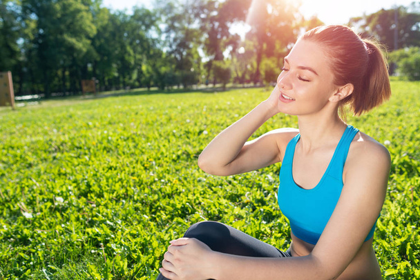 Beautiful smiling girl in sportswear relax in park. Young woman with closed eyes sitting on green grass. Training and meditation outdoor at sunny summer day. Morning exercises and healthy lifestyle - Fotografie, Obrázek